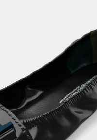 Gros plan d'une chaussure plate en cuir verni noir avec un bord élastique et une boucle décorative bleue sur le bout.