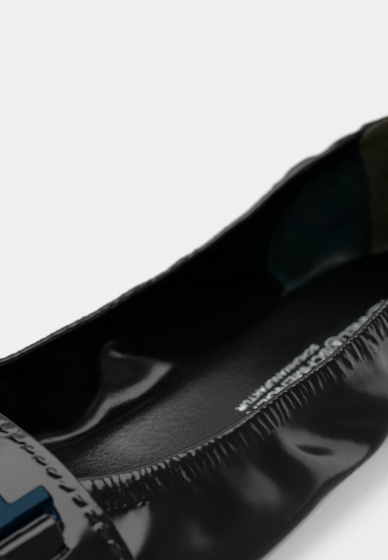 Gros plan d'une chaussure plate en cuir verni noir avec un bord élastique et une boucle décorative bleue sur le bout.