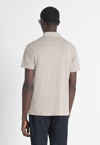Camiseta de polo de manga corta beige con un cuello clásico, textura suave y aberturas laterales, combinada con pantalones oscuros y gafas.