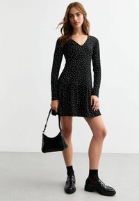 Vestido negro de manga larga con lunares blancos, escote en V, cuerpo ajustado, falda acampanada, combinado con un bolso negro y mocasines brillantes.