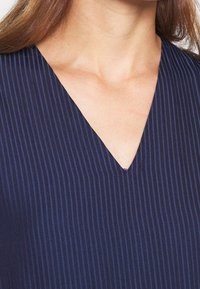 Námořnická V-čková blůza s vertikálními pruhy, vyrobená z hladkého materiálu s jemným leskem. Výstřih je ostrý a dobře definovaný.