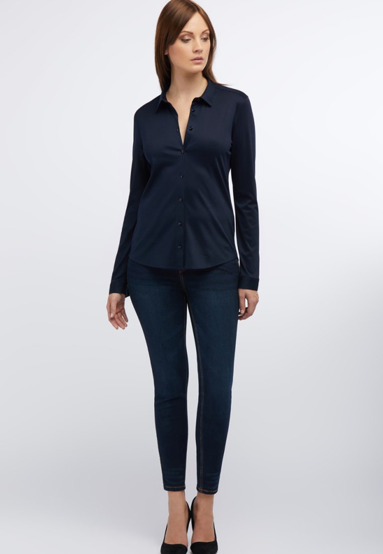 Navy long-sleeve button-up shirt with a collar, paired with dark skinny jeans and black heels. Smooth fabric with subtle sheen.