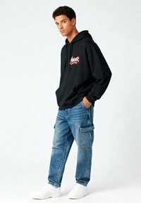 Felpa nera con cappuccio e logo rosso, abbinata a jeans cargo azzurri chiari con tasche laterali e sneakers bianche. Outfit casual.
