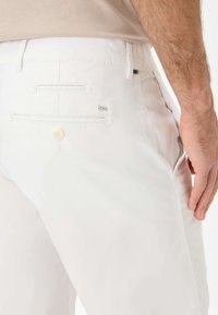 White cotton pants featuring two back pockets with button closures, a small logo tag, and a smooth texture. Tailored fit design.