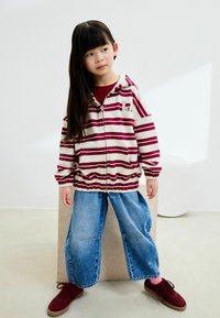 Sweat à capuche zippé rayé en crème et bordeaux, associé à un pantalon en denim à jambes larges. Des baskets rouges complètent la tenue. Look simple et décontracté.