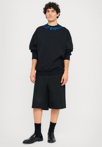 Sweatshirt preto com estampa gráfica azul na gola, combinado com calções pretos. Conjunto finalizado com meias e sapatos pretos.
