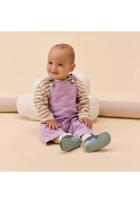 Un bambino sorridente seduto su un pavimento beige, che indossa una tuta viola, una maglia a maniche lunghe a righe e delle scarpe blu, con un morbido cuscino bianco dietro di lui.