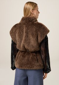 Bruine nep-bont gilet met een hoge kraag, losse pasvorm en een ceintuur in de taille. Onder het gilet zijn doorschijnende zwarte kanten mouwen zichtbaar.