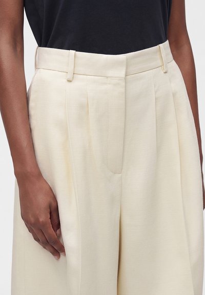 Personne portant un pantalon plissé crème taille haute avec passants de ceinture et un haut noir rentré, main reposant sur le côté.