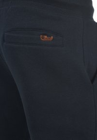 Navy sweatpants feature a ribbed waistband, a small tan leather logo patch, and a back pocket with a horizontal opening. Smooth texture.