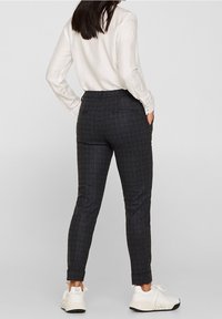 Vrouw in donkergrijze geblokte slim fit broek, witte blouse en witte sneakers, staande met handen in de zakken, met de rug naar voren gericht.