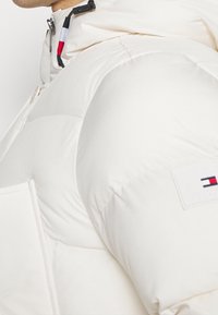 Hvid pufferjakke med hætte, sidelommer og et branding patch med rød, hvid og blå kant ved lynlåsen og kraven.