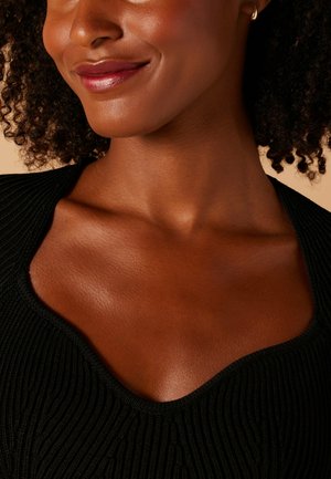 Gros plan sur un vêtement noir texturé avec un large décolleté, mettant en avant un teint lisse et une couleur de lèvres naturelle. Des détails subtils incluent un tissu côtelé.