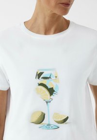 Weißes T-Shirt mit einem grafischen Druck eines Cocktailglases mit Zitronenscheiben und Blättern, in lebendigen Farben und mit glatter Textur.