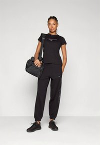 Svart atletisk antrekk med en tettsittende t-skjorte og løse bukser, begge med en logo, kombinert med svarte sko og en svart veske. Jevn tekstur.