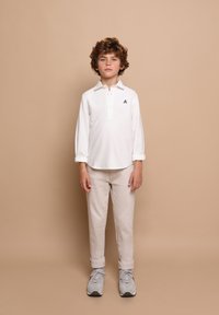 Weißes Langarm-Polo mit einem Knopfkragen, schwarzem Emblem auf der Brust, kombiniert mit beigen Hosen und grauen Sneakern, vor einem neutralen Hintergrund.