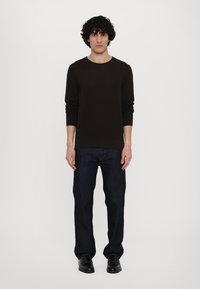 Dunkelbrauner Strickpullover mit langen Ärmeln, kombiniert mit dunkelblauen geraden Jeans und schwarzen Schuhen, getragen von einem stehenden männlichen Model.