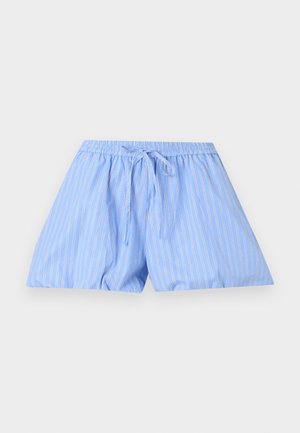 Femme debout portant une chemise rayée bleu clair à manches longues avec boutons et un short assorti, ainsi que des chaussures Mary Jane noires sur fond blanc.