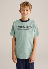 Zelené tričko s krátkym rukávom a námořníckym golierom. Na prednej strane je vytlačený text "TEAM NO SLEEP" a súradnice. Jemná textúra látky.