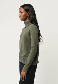 Olivgrüner Rollkragenpullover aus weichem Stoff, mit langen Ärmeln und geripptem Saum. Trägt er zu schwarzen Hosen, seitlich präsentiert.