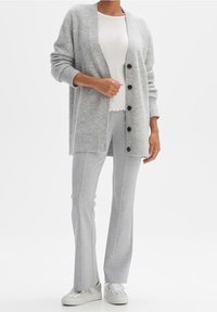 Šedý pletený kardigan s velkými černými knoflíky, nošený přes bílý top a šedé slim fit kalhoty. Má volný střih a žebrované manžety. Celý outfit doplňují tenisky.