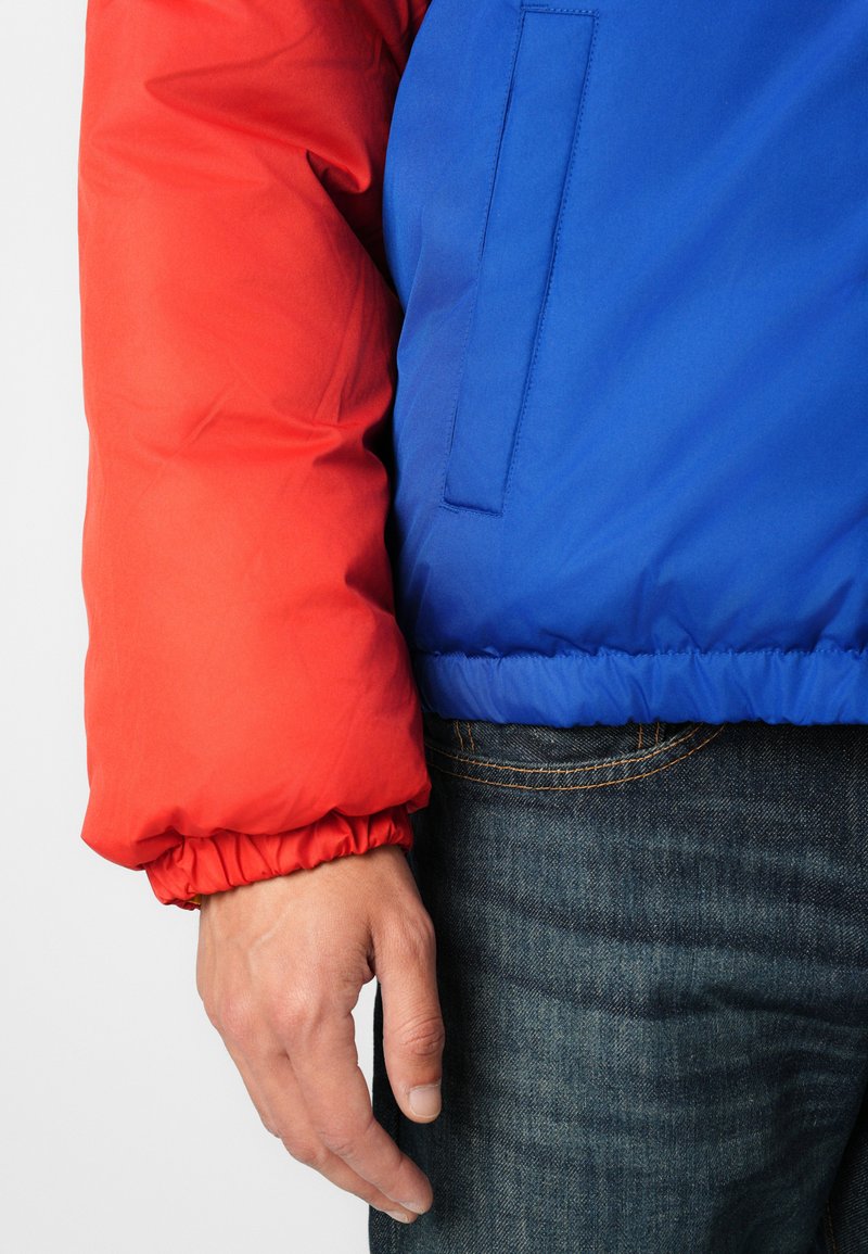 Zbliżenie na rękę w pobliżu rękawa czerwonej kurtki puchowej i niebieskiej części kurtki, noszonej z ciemnymi dżinsami.
