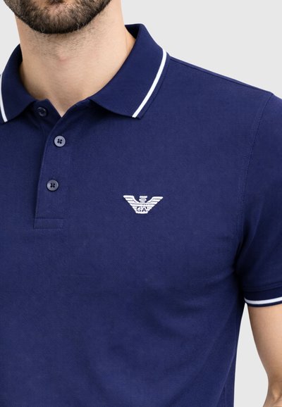 Άνδρας που φοράει μπλε σκούρο πόλο μπλουζάκι με λευκή λεπτομέρεια στο γιακά και στα μανίκια, με μικρό λευκό κεντητό λογότυπο αετού στο στήθος.
