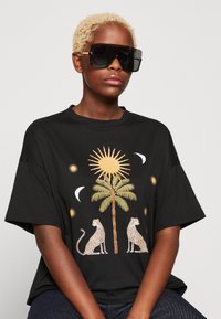 T-shirt oversize nera con una stampa grafica di due leopardi, una palma, un sole e lune crescenti in toni dorati e beige.