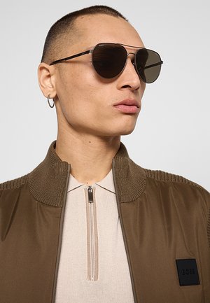 Uomo con capelli corti che indossa occhiali da sole aviator scuri, una giacca marrone con colletto a coste e una camicia beige con zip sotto.