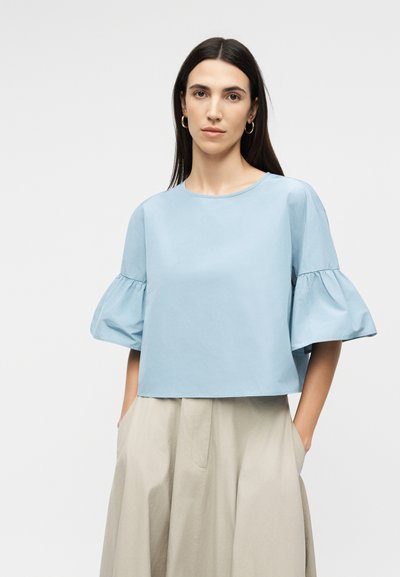 Femme aux longs cheveux foncés portant des boucles d'oreilles en or en forme de créoles, un chemisier bleu clair à manches bouffantes et un pantalon beige taille haute, les mains dans les poches.