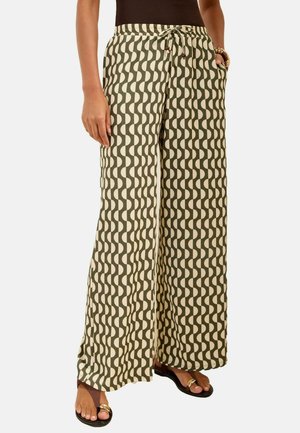 Woman wearing high-waist wide-leg pants with green and beige wavy pattern, black top, sandals, and bracelets on left wrist.