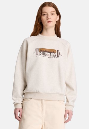 Ung kvinde med langt rødt hår iført en beige Timberland sweatshirt og lyse fløjlsbukser mod en ensfarvet baggrund.