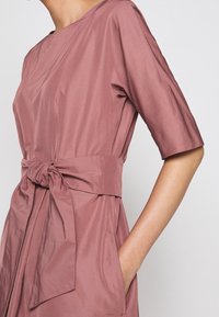 Růžové šaty z hladké tkaniny, s kulatým výstřihem, krátkými rukávy, detaily vázání v pase a bočními kapsami.