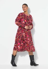 Langärmliges, knielanges Kleid mit einem wirbelnden Muster in Rosa, Rot und Orange. Kombiniert mit schwarzen Cowboystiefeln steht das Modell vor einem weißen Hintergrund.
