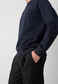 Námořnický crewneck svetr vyrobený z hladkého materiálu, se malým logem na hrudi a žebrovanými manžetami, v kombinaci s černými kalhotami.