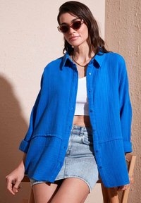 Lyst blå skjorte med strukturstof, kombineret med en tætsiddende hvid tanktop og denimnederdel. Solbriller fremhæver outfittet.