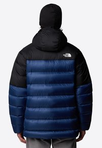 Kurtka puchowa z czarnym kapturem i górą, a dolna część jest niebieska, pikowana. Wyróżniający się błyszczący materiał oraz logo North Face na plecach.