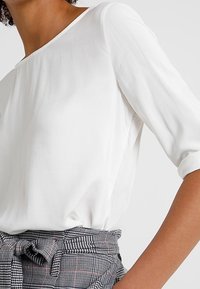 Kvinna iklädd en vit långärmad blus instoppad i högmidjade grå rutiga byxor med ett knutet tygbälte.