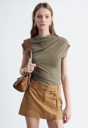 Kvinde iført olivengrøn ærmeløs top og beige nederdel, der bærer en brun lædertaske over skulderen, mod en ensfarvet lys baggrund.
