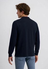 Mann mit lockigem braunem Haar, der ein navyblaues Langarmshirt und hellblaue Jeans trägt, steht mit dem Rücken zu einem schlichten weißen Hintergrund.