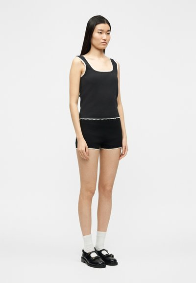 Femme debout portant un haut noir sans manches et un short assorti avec un bord blanc, des chaussettes blanches, et des mocassins noirs sur un fond uni.