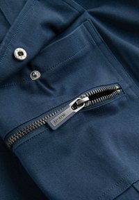 Gros plan d’un tissu bleu avec une fermeture éclair en métal et deux boutons-pression en métal sur une poche de vêtement.