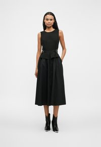 Μαύρο φόρεμα με εφαρμοστό τοπ, μέση peplum και ροή, μεσαίου μήκους φούστα. Περιέχει μεταλλικά ασημί αξεσουάρ και τσέπες.