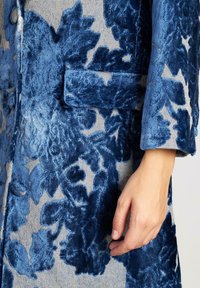Abrigo de terciopelo azul con estampado floral en relieve; presenta una mano visible en la manga y un bolsillo en el pecho.