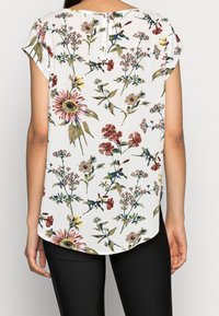 Kvinde iført hvid kortærmet bluse med flerfarvet blomsterprint og sorte bukser, vist bagfra.