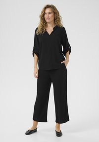 Blusa nera con scollatura a V e maniche arrotolate a tre quarti, abbinata a pantaloni neri larghi e scarpe nere basse, che presenta una silhouette elegante.