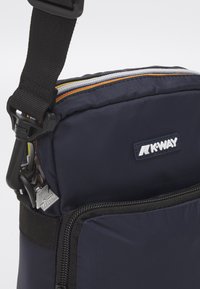 Marineblaue K-Way Umhängetasche mit schwarzem verstellbarem Riemen, mehreren Reißverschlussfächern und sichtbaren K-Way-Logoaufnähern.