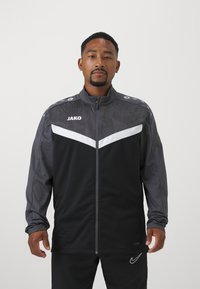 Zwarte en grijze sportjacket met een volledige rits, chevron witte streep en logo op de borst. Voorzien van schouderaccenten en textuurstof.