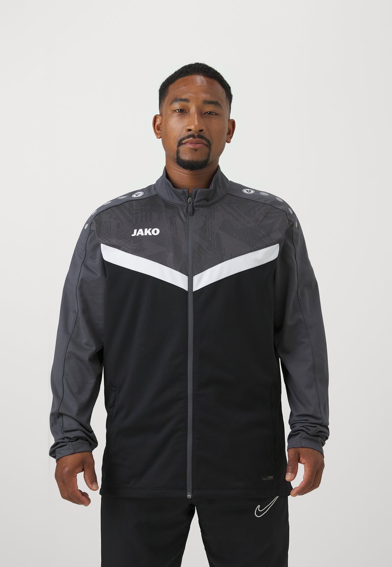 Zwarte en grijze sportjacket met een volledige rits, chevron witte streep en logo op de borst. Voorzien van schouderaccenten en textuurstof.