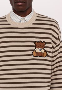 Beige és barna csíkos pulóver, bordás kerek nyakkal, amelyen egy hímezett teddy mackós folt található a bal mellen.